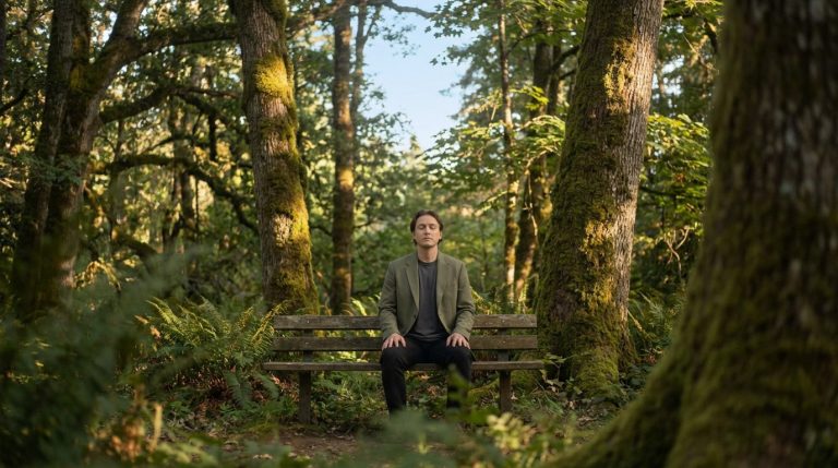Un homme en veste verte médite, les yeux fermés, sur un banc en bois dans une forêt luxuriante et ensoleillée. Ambiance paisible.